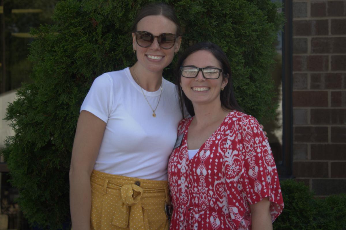 Two women stand close together, with one arm around the other. The woman on the left is wearing sunglasses, with her hair styled in a tight, middle-parted updo. She’s dressed in a white t-shirt and a yellow skirt. The woman on the right has her dark hair down and is wearing prescription glasses. She also has a necklace around her neck and is dressed in a loose-fitting red top with white patterns.