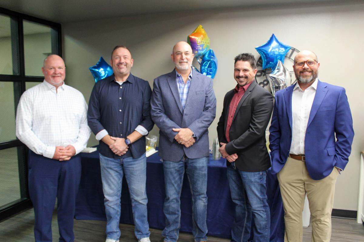 Five people stand in a row in front of a table with balloons behind them.