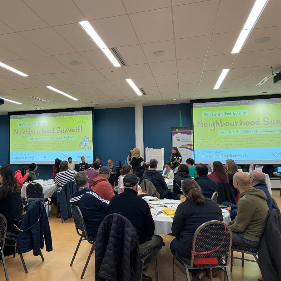 Group of people sitting at round tables looking at screen that reads neighbourhood summit