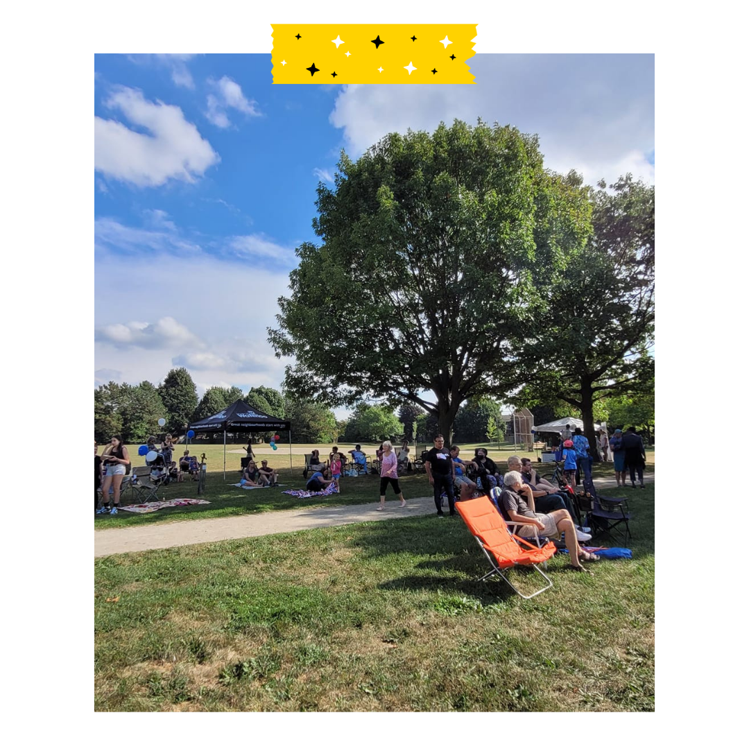 Image of people outside sitting under trees and tents in a park on a sunny day.