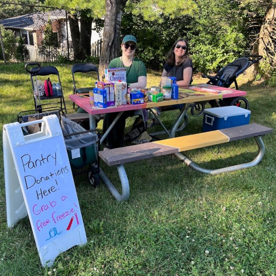 2 people sitting at a picnic table, with sign requesting food donations