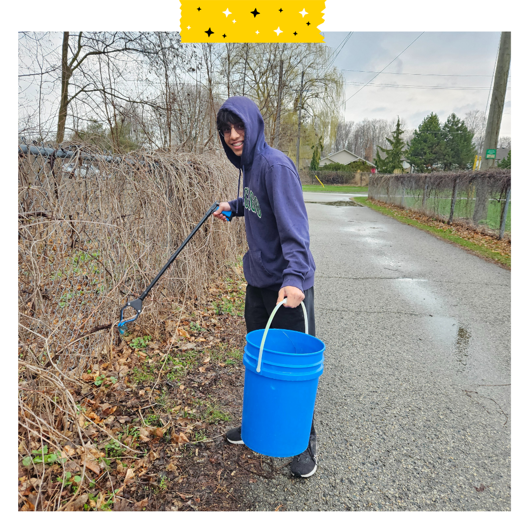 A person with a dark blue hoodie on, with a garbage picker and a blue bucket picking up garbage on a trail. 