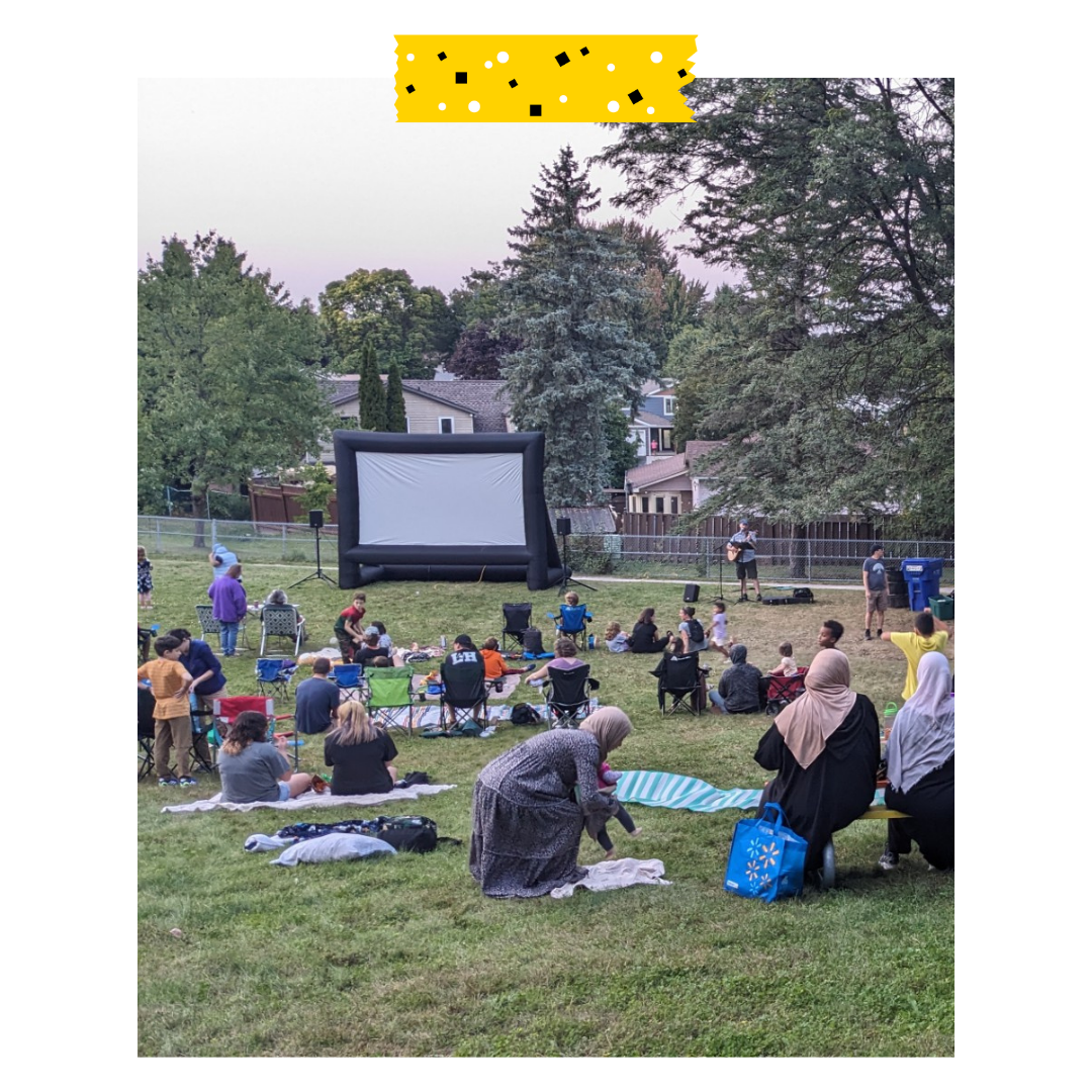 Neighbours gather on the grass with blankets and folding chairs in front of a movie screen