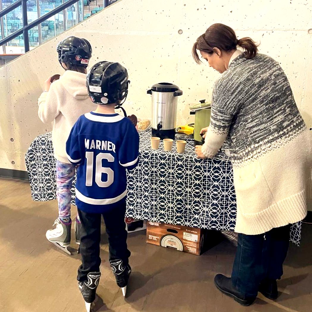 two kids in wearing helmets and skating gear by a table with drinks and snacks. One adult pouring drink from table. 