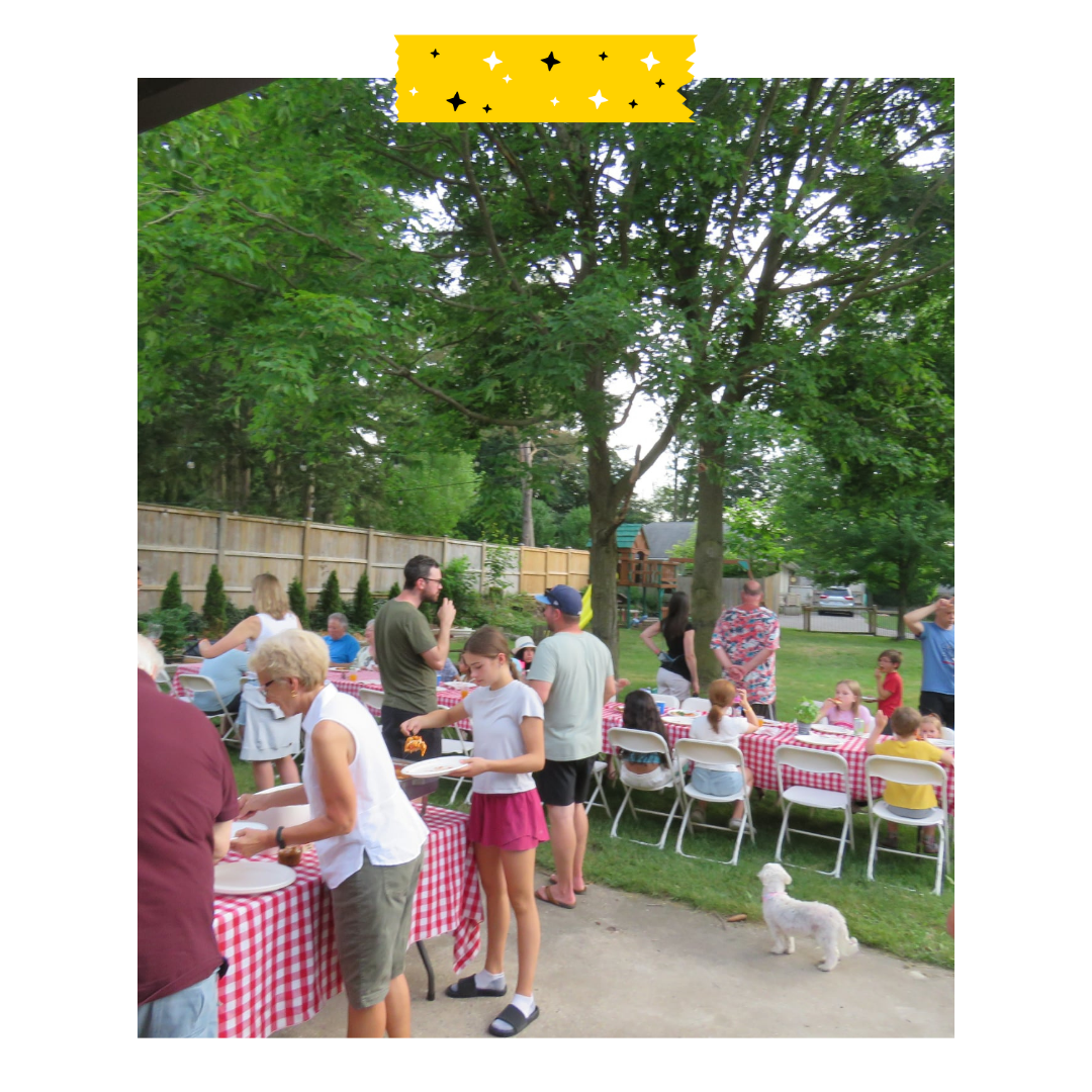 Neighbours gathered around tables and outside. There is food and drinks on the tables with picnic table cloths.
