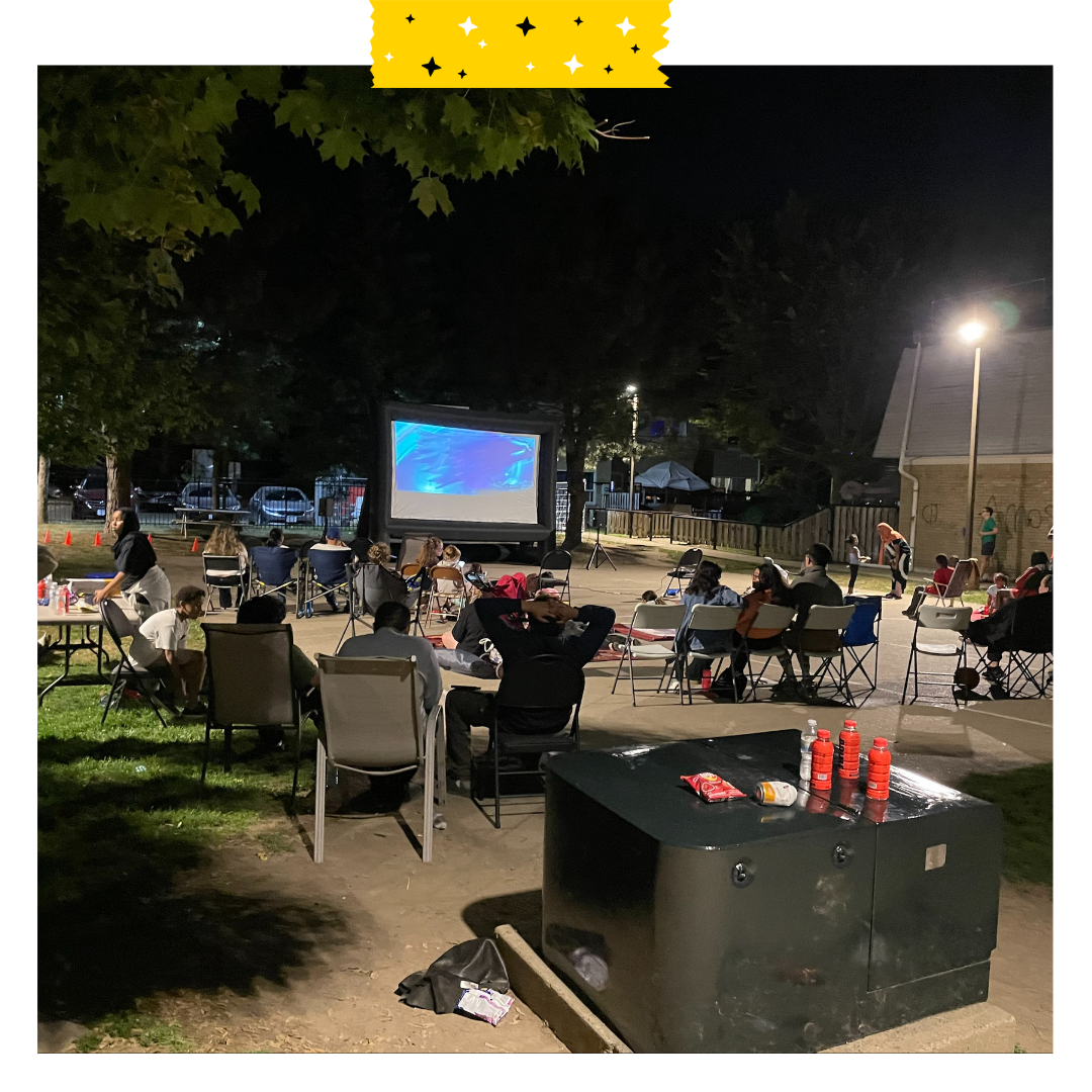 Outdoor movie set up, people sitting in chairs outside on pavement looking at the screen in the dark.