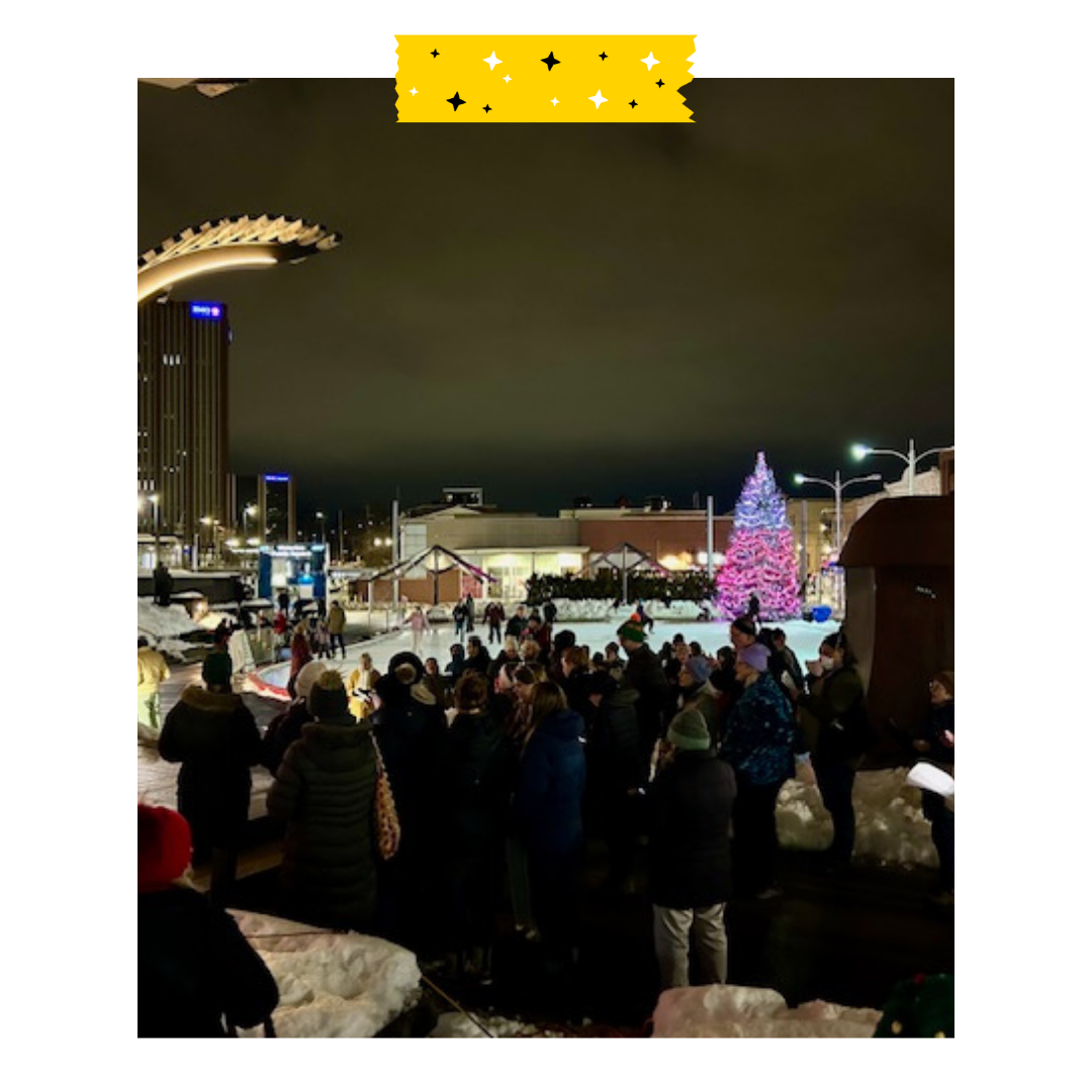 A group of people standing outside in the dark with lights on in the background.