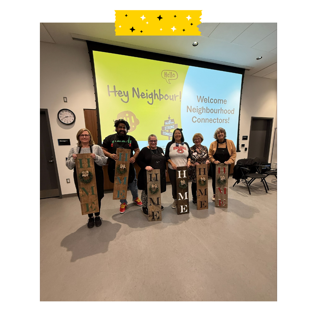 6 people standing holding a home welcome sign in front of a backdrop that says hey neighbours welcome neighbourhood connectors.