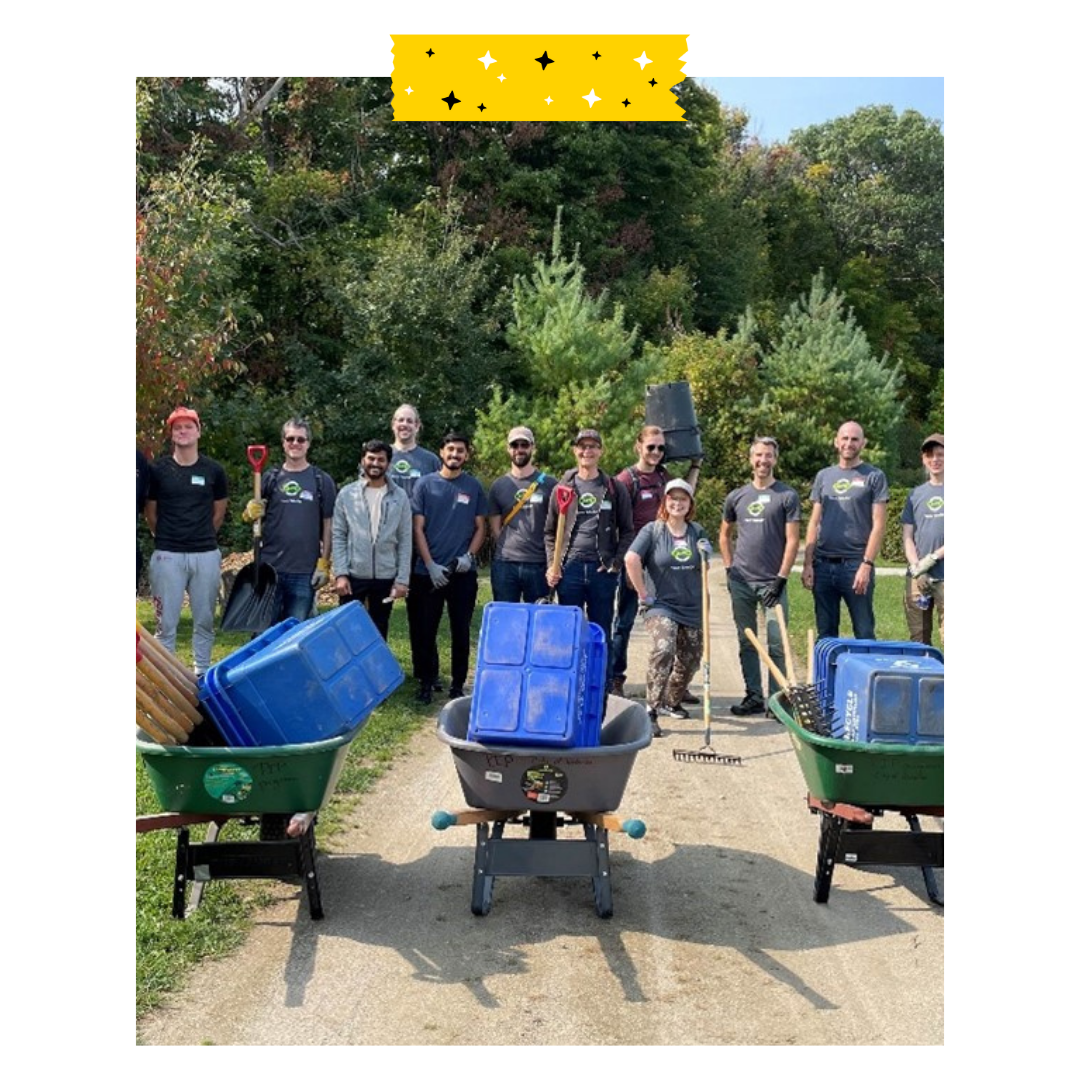 People are standing outside in a park, posing for a photo with wheelbarrows in front.