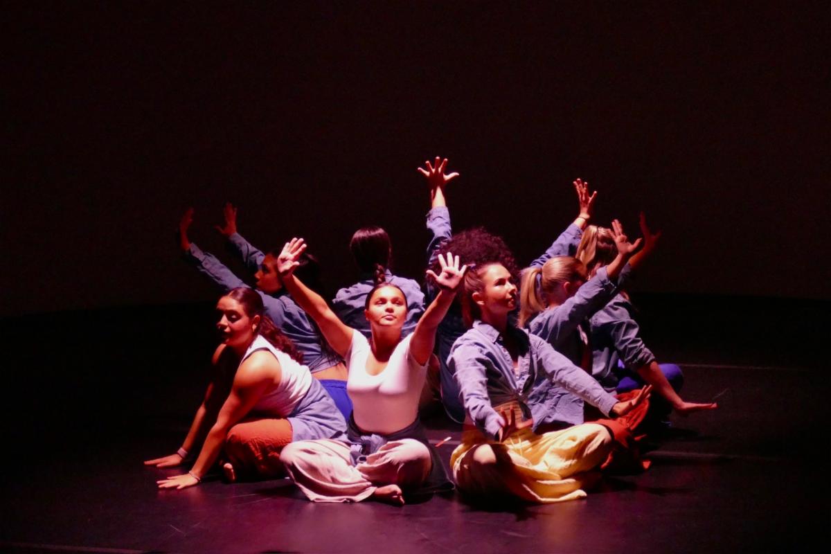large group of dancers seated on the stage floor front-facing in a circle with their hands up