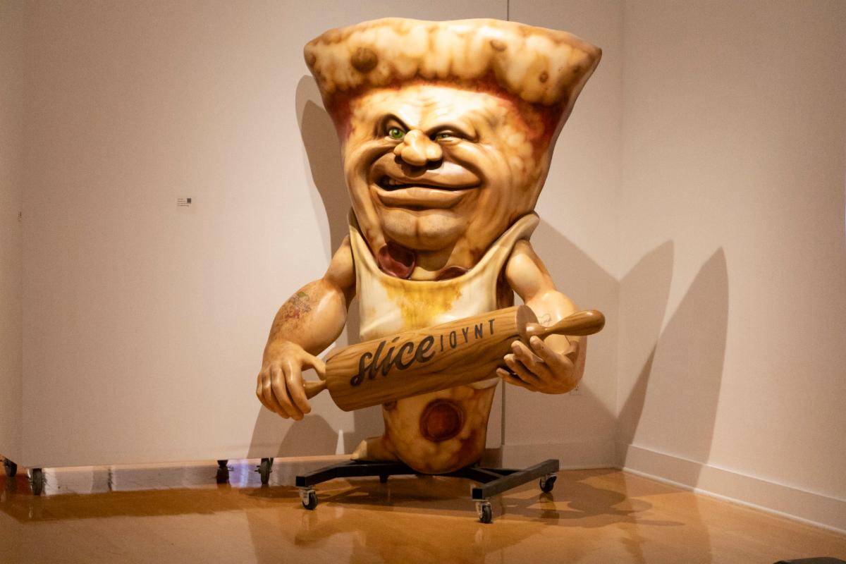 sculpture of a large piece of pizza with a smirking old man's face holding a rolling pinin the art gallery