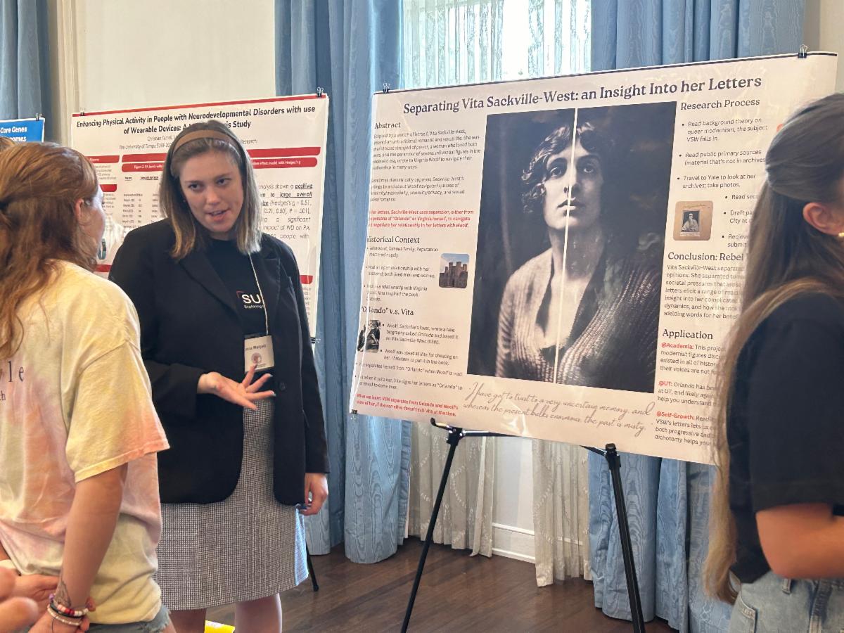 Lena Malpeli standing in front of a research poster and talking to guests about it