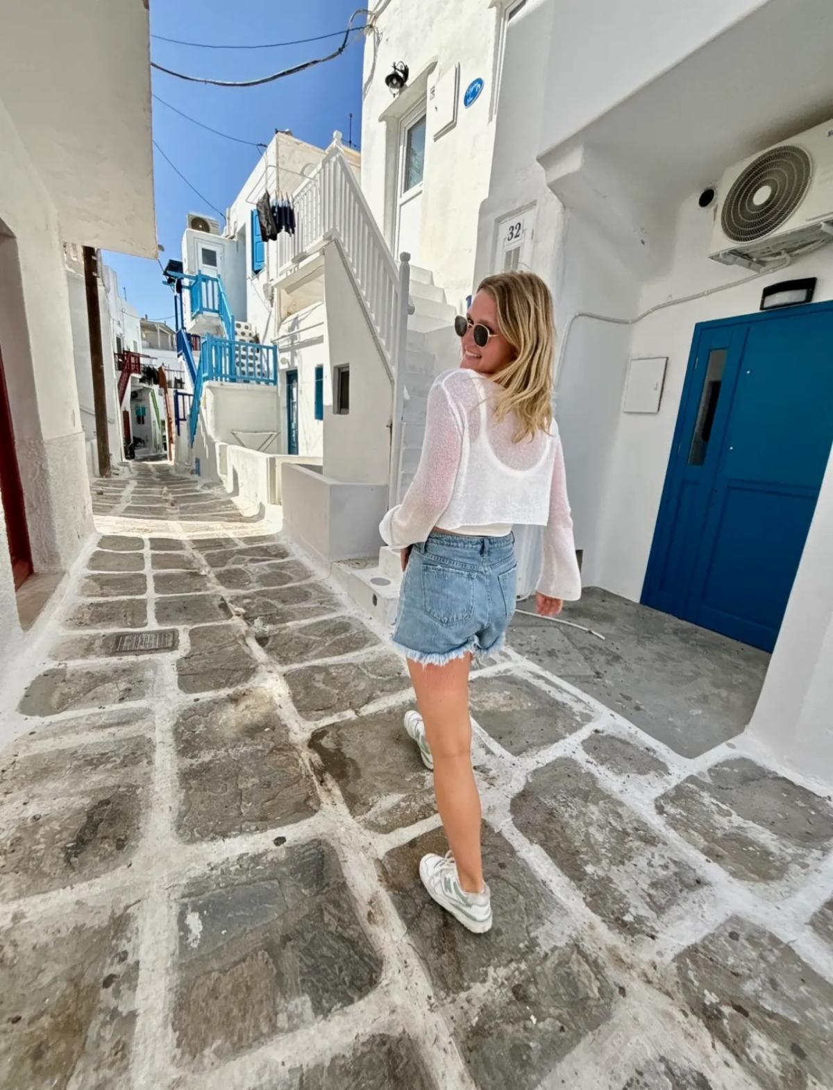 Libby in causal wear walking in a neighborhood ally in Greece with white homes and buildings