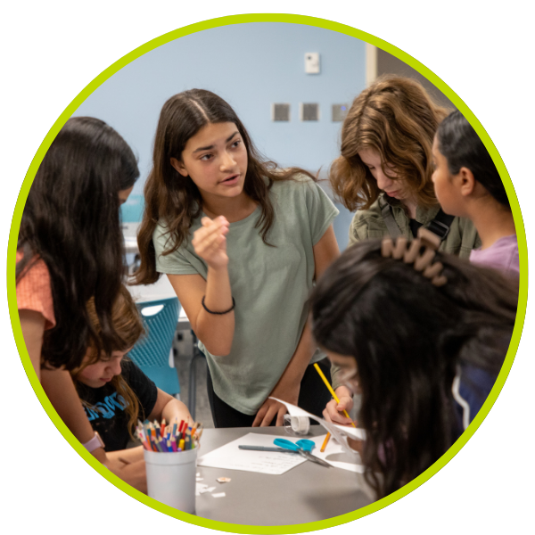 Five adolescent girl standing around a table and one of them leading a discussion.