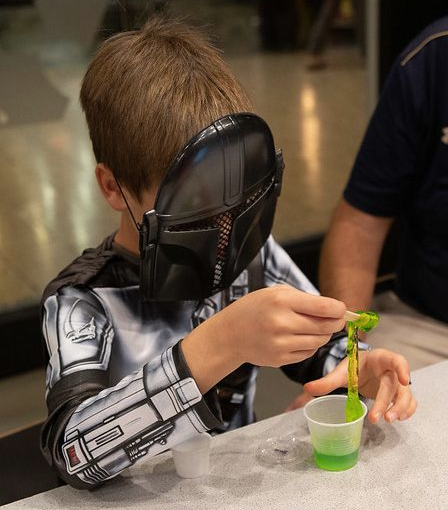 A young boy dressed as a star wars character (complete with mask) sits at the Experiment Bar counter. A cup of bright lime green slime is in front of him, and he's using a popsicle stick in one hand to lift some out of the cup and let it ooze back down.  An adult seated next to him is partly visible in the background.