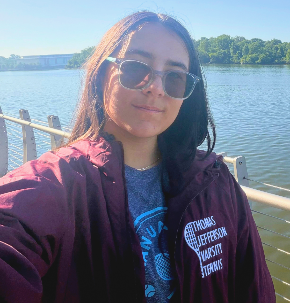 A high school girl with long dark hair and sunglasses on poses for a selfie overlooking water. She's wearing a dark maroon sweatshirt that says Thomas Jefferson Varsity Tennis and smiling slightly.
