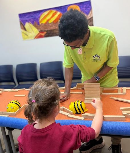 A high school boy in a green Lab volunteer shirt stands behind a table, guiding a elementary schooler through an activity. A maze made of wooden Keva blocks is on the table between them, and a palm-sized robot shaped like a computer mouse is making its way through. Another robot has crashed into the side of the maze further down the table. Behind them, a large color drawing of Jupiter and its moons is displayed on the wall. 