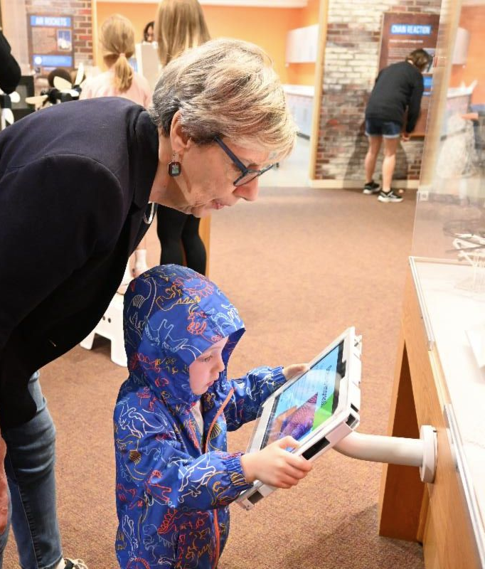 A young boy (4-6 yrs) in a dinosaur-patterned blue coat examines the screen attached to a 3D printer.  The screen sticks out of the printer housing on a tube, and the boy has one hand on either side of it. On the screen, a box of bright colors is visible on one side. Behind the young boy, an older woman with dark glasses leans over to look at the same thing.