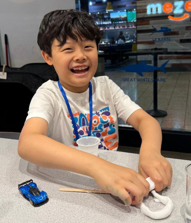 A young boy in late elementary school laughs at the camera from his seat at the Experiment Bar. On the counter in front of him is a small pile of white slime, which he is merging into a tube with his hands. He is clearly delighted.