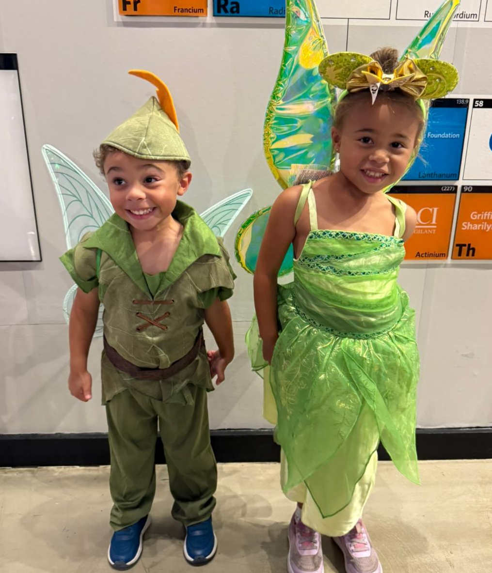 Two small children (roughly three and five) pose for the camera in their Halloween costumes. They are both dressed as fairies, all in lime green with wings on their backs. 