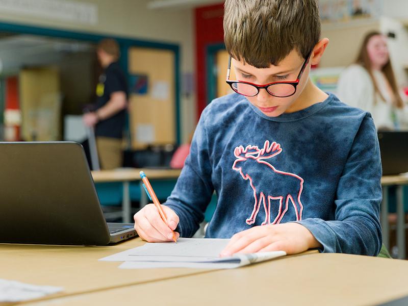 A boy works in a classroom next to a laptop