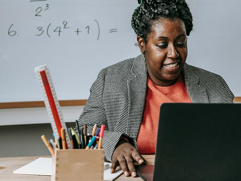 A teacher looks at a laptop in front of a whiteboard with math equations