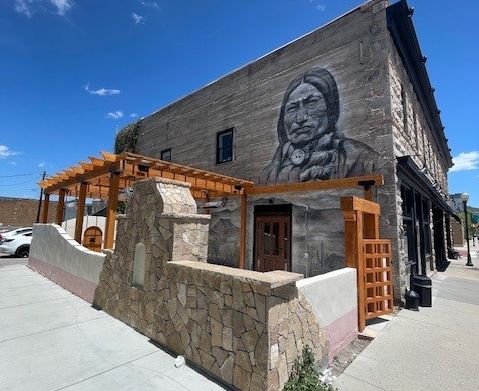 Exterior photo of the Next Door Bar patio with stucco low wall with stone fireplace, wood pergola and Native American portrait mural painted on exterior wall of the building