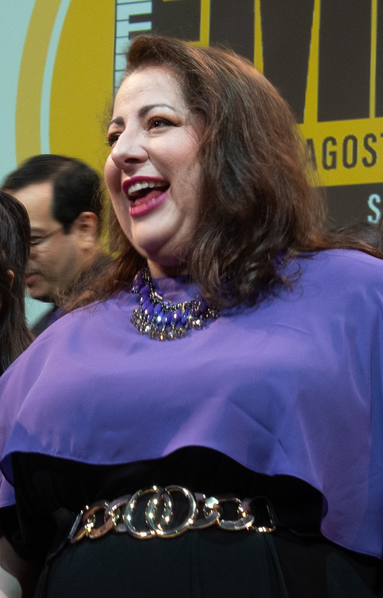 Michelle is smiling on the Met Stage with the FMDG logo behind her. She is wearing a purple caplet over a black dress.