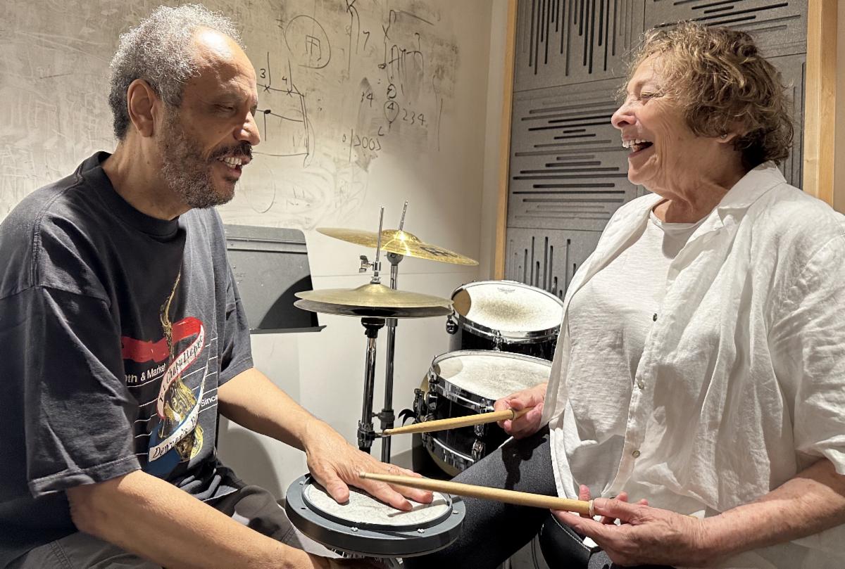 Barbara and Vanderlei are sitting on either side of a drum. Barbara has drum sticks in her hands. Vanderlei has his hand on the drum. Barbara is wearing a white top. Vanderlei is wearing a black tshirt. 