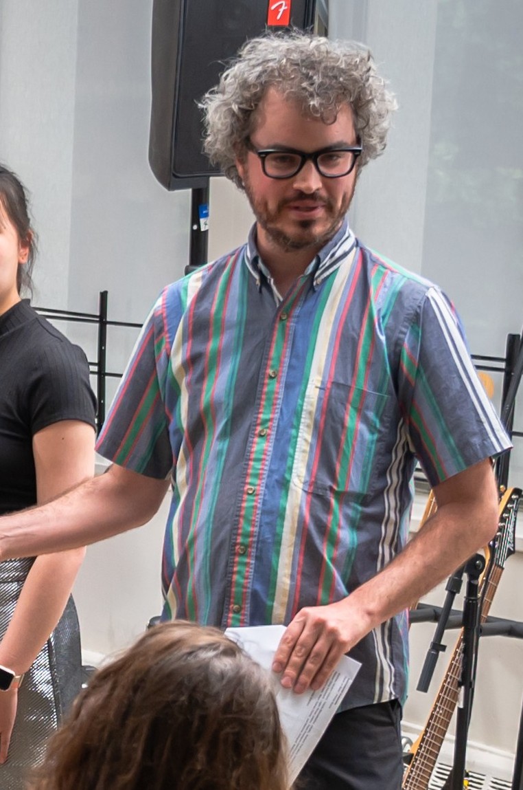 Anthony Vine speaking to an audience and gesturing with his hand. He is wearing a vertically striped short-sleeved collared shirt. The stripes are white, blue, red, and green in varying widths.