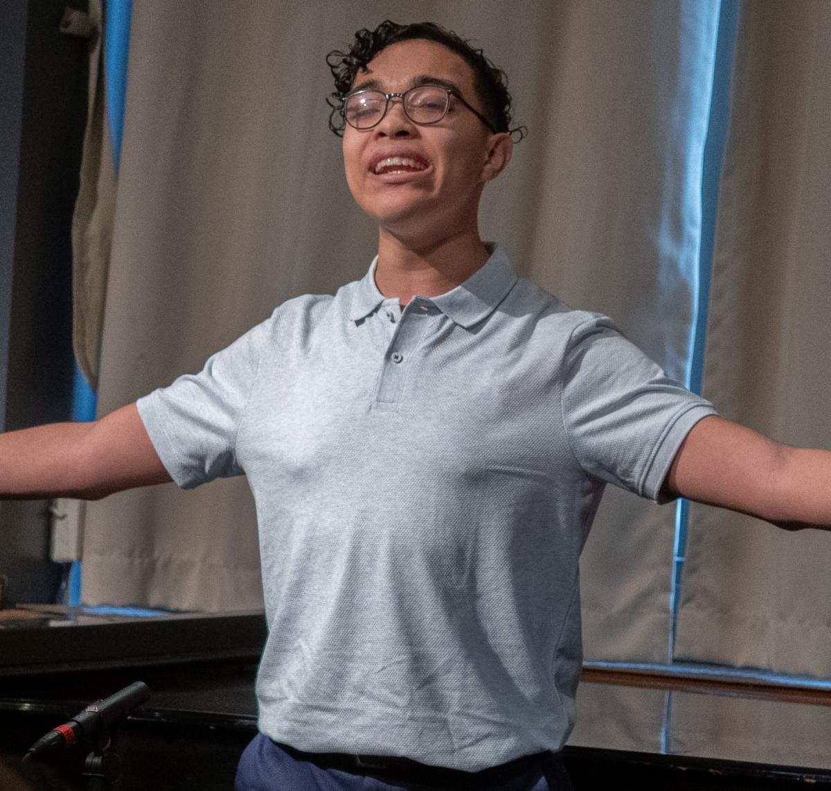 Matthew Herrera is singing with his arms outstretched and smiling. Matthew is wearing a grey short sleeved collared shirt. He has short black hair and wears glasses.