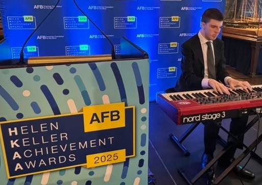 Logan Riman playing a keyboard in front a blue AFB banner. To Logan's right there is a sign which reads Helen Keller Achievement Awards 2025. Logan is wearing a dark suit and tie with a white collard shirt.
