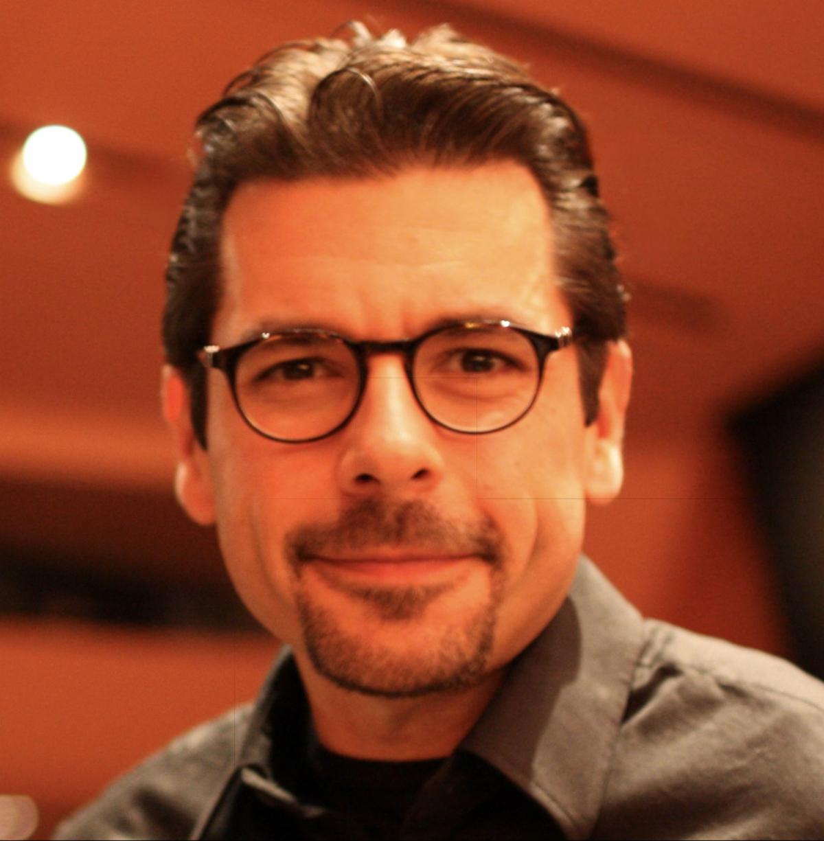 A headshot of Rob. He has short black hair and a black goatee. Rob is wearing black glasses and a grey collared shirt. 