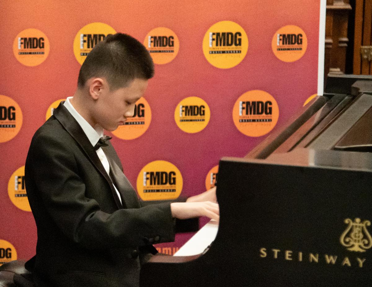 Hoawen is playing the piano dressed in a black tuxedo and bow tie.