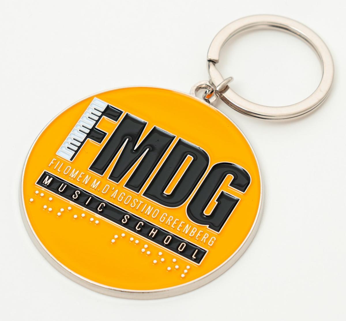 An FMDG Music School Keychain. The keychain has the schools logo printed in black and white on a gold background. The words music school are printed in raised braille.