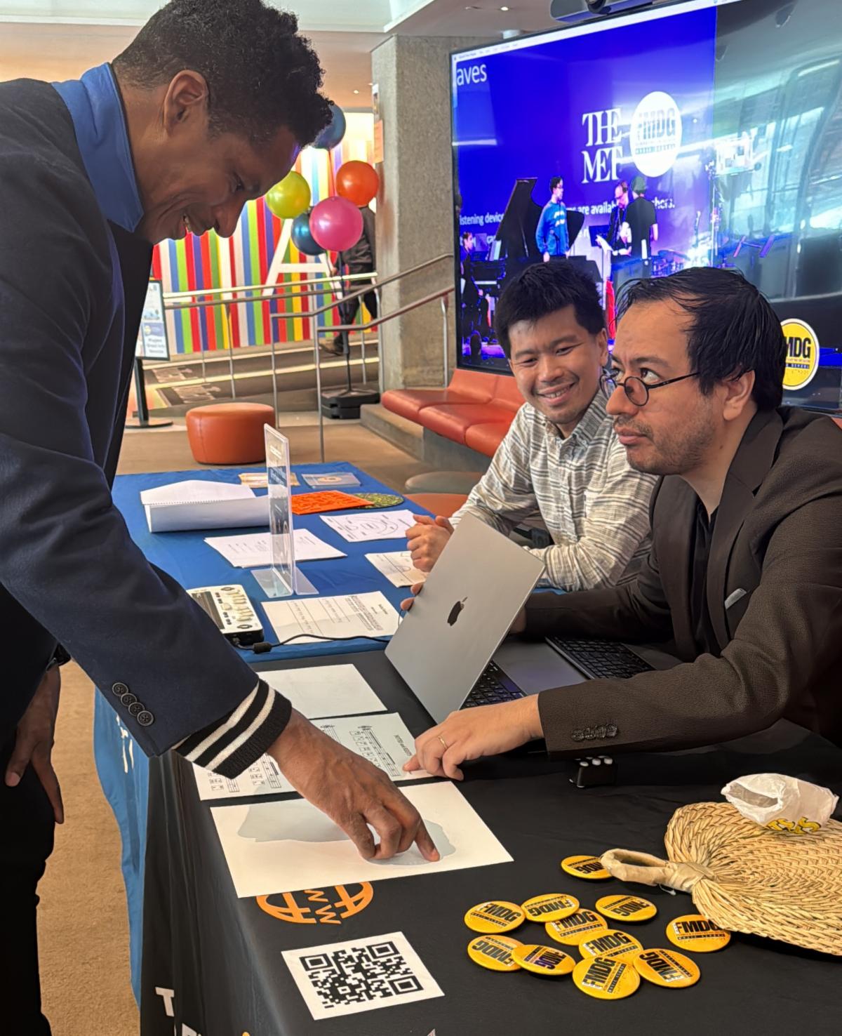 Pablo and Richard sit as a table on which there are FMDG buttons a laptop and printed information in ink and braille. They are engaged with a visitor to the table. 