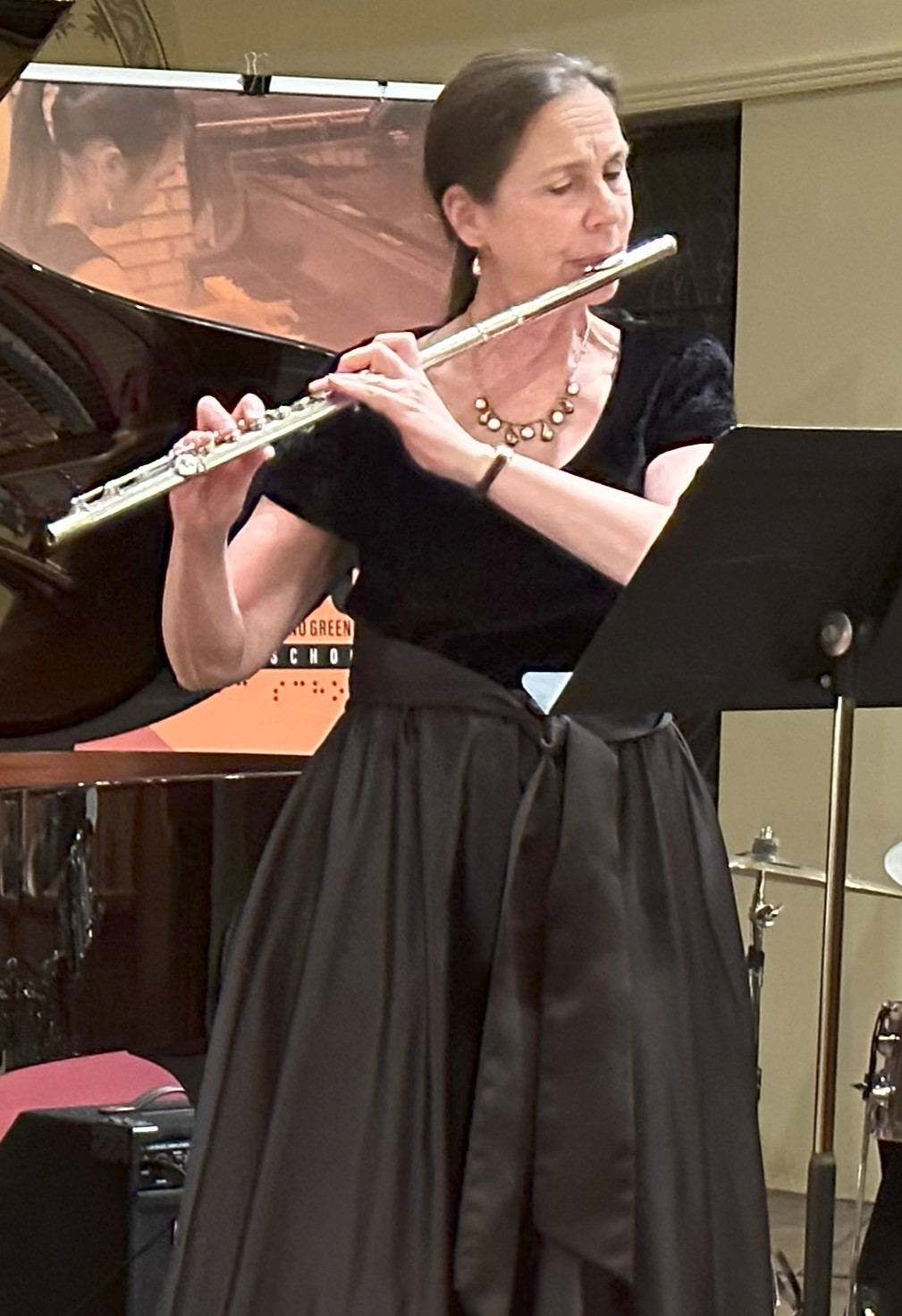 Katharine is standing and playing the flute. She is wearing a long black dress with short sleeves and a shiny necklace. Her long brown hair is pulled back in a pony tail. 