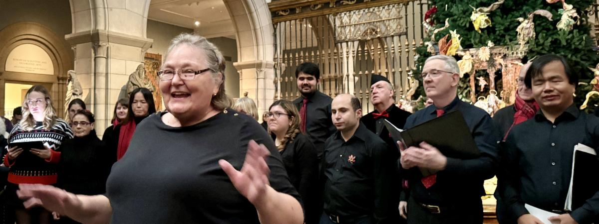 A close up of Dalia in front of a section of the adult chorus with a Christmas tree behind them. The chorus is wearing black with splashes of red. Dalia has an exuberant expression.