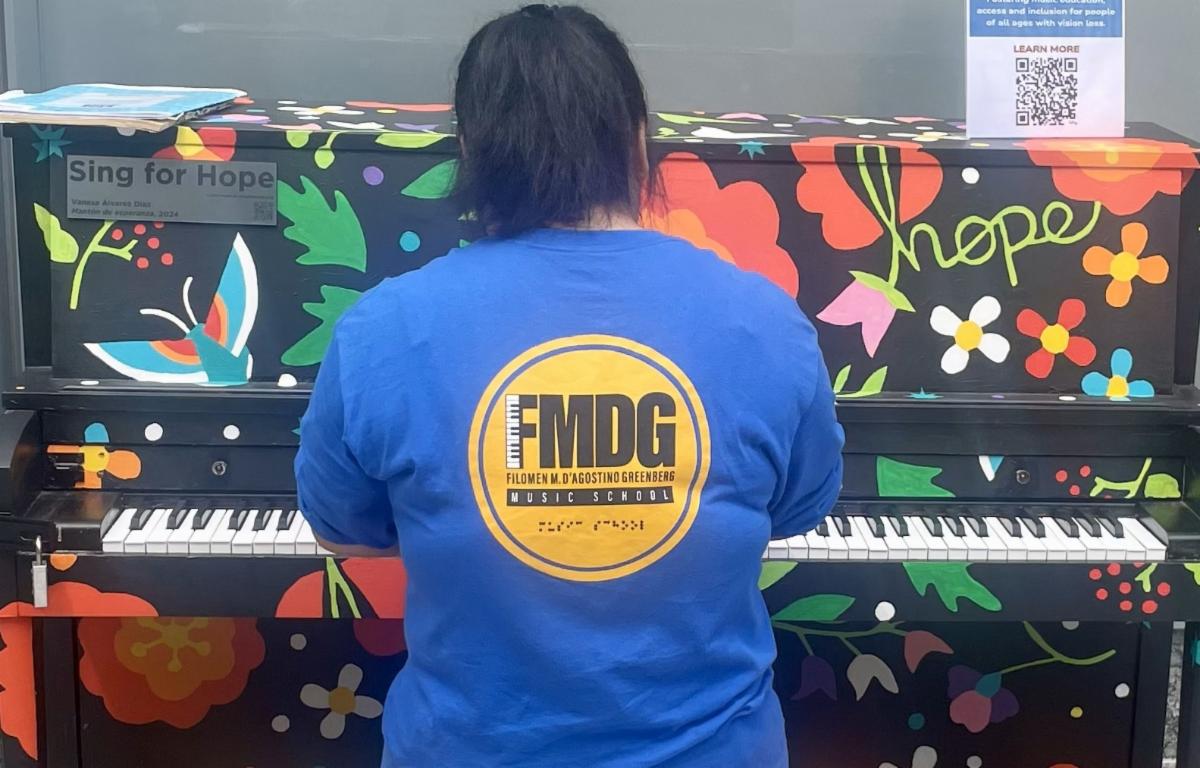 A young piano player sits at a colorfully decorated piano with their back to the camera. The pianist is wearing a blue long sleeve tshirt with the FMDG logo in gold and black. The piano is decorated with flowers butterflies leaves and berries.