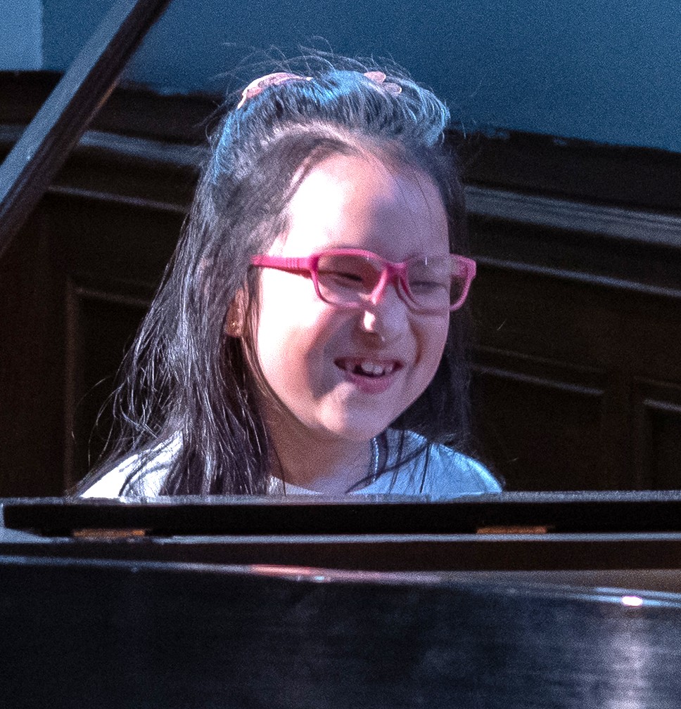 Chenyu is playing the piano. She is facing the camera and smiling. Chenyu has long black hair and is wearing pink glasses.