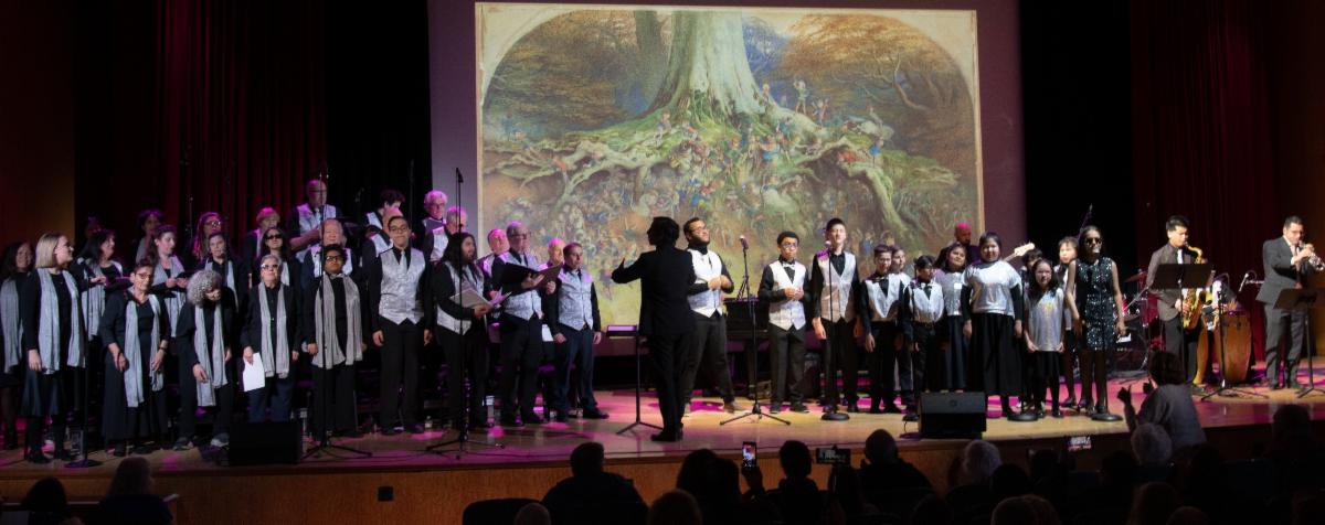 The adult and youth choruses on stage at the Met. The signature image of the Enchanted Tree is projected behind them.