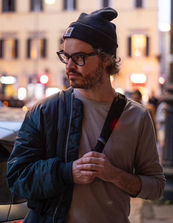Anthony Vine walking on a city street. He is wearing a black jacket over one shoulder over a grey collarless shirt. He has big black glasses and a black stocking cap on his head. He is looking away from the camera.