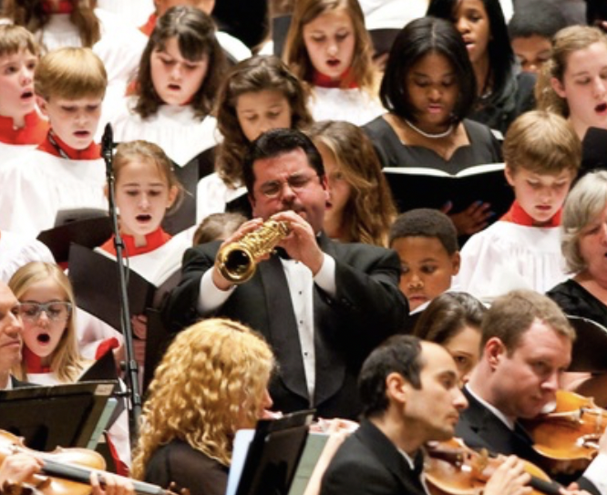 Rob playing soprano saxophone among a chorus of young people and an adult orchestra. Rob is wearing formal concert attire.