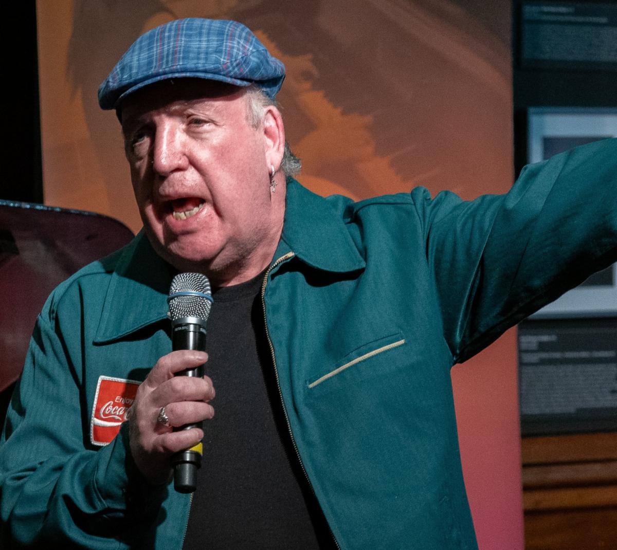 Raymond Lonergan is singing holding a mic and one arm outstretched. Ray is wearing a blue and green tartan cap and a dark green jacket over a black tshirt.