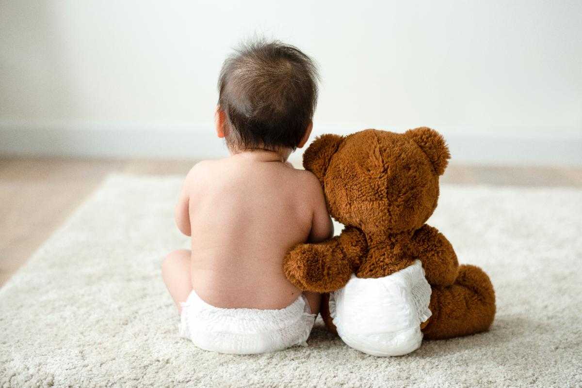 Dark haired baby wearing only a diaper sits on a carpeted floor with his back to the camera.  Beside him sits a brown stuffed, teddy bear also wearing a diaper with his back to the camera.  The bear has his left arm gently touching the back of the baby.