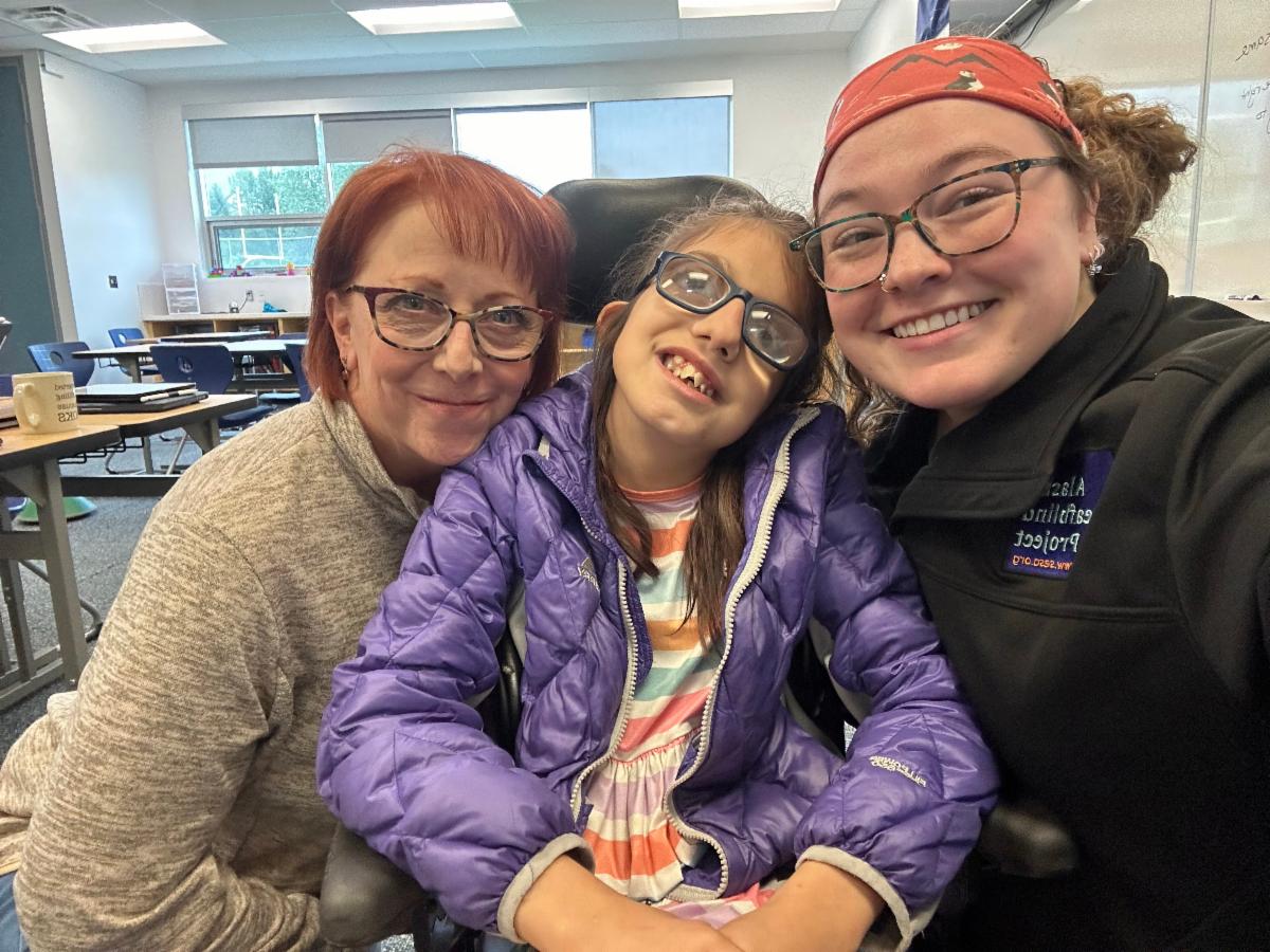 An Adult woman with red hair rests her head on the left shoulder of a girl wearing glasses in a purple jacket. An Adult woman in glasses with an orange headband smiles over the right shoulder of the girl