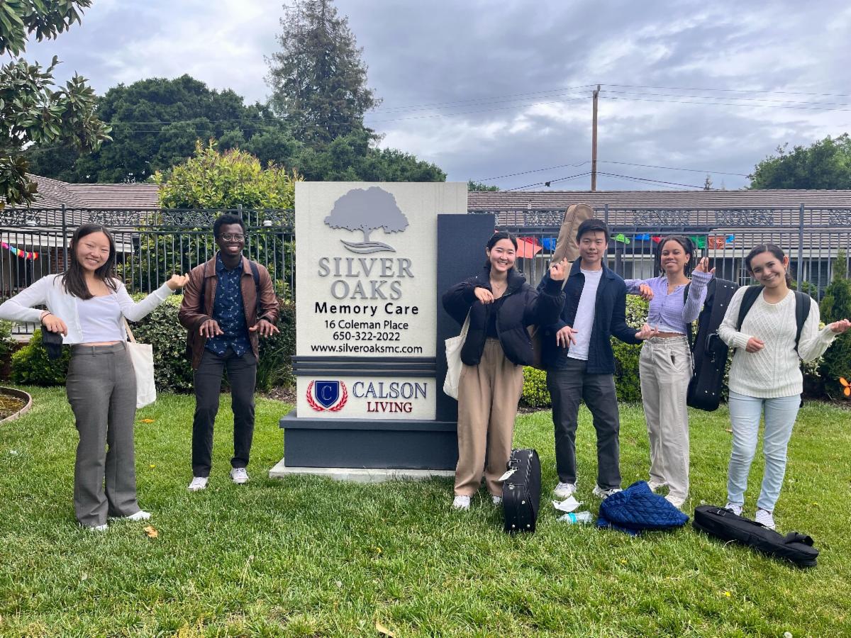 Students pose pretending to play instruments by Silver Oaks Memory Care sign