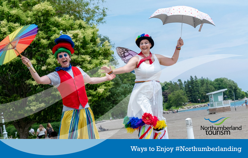 Two stilt walkers holding hands and parasols walking along Cobourg beach. Photo by Peg McCarthy