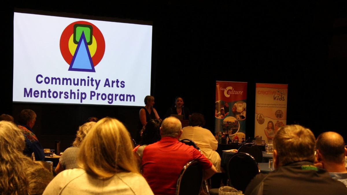 A photo the crowd at the 2024 AGM, with the logo of the Community Art Mentorship Program in the background.