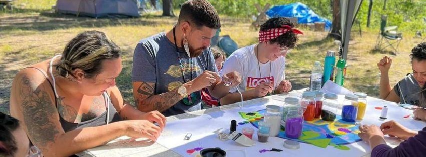 Participants and Knowledge Keepers from this year’s OUT on the Land camp sitting around table beading. The background shows trees and camping tents.